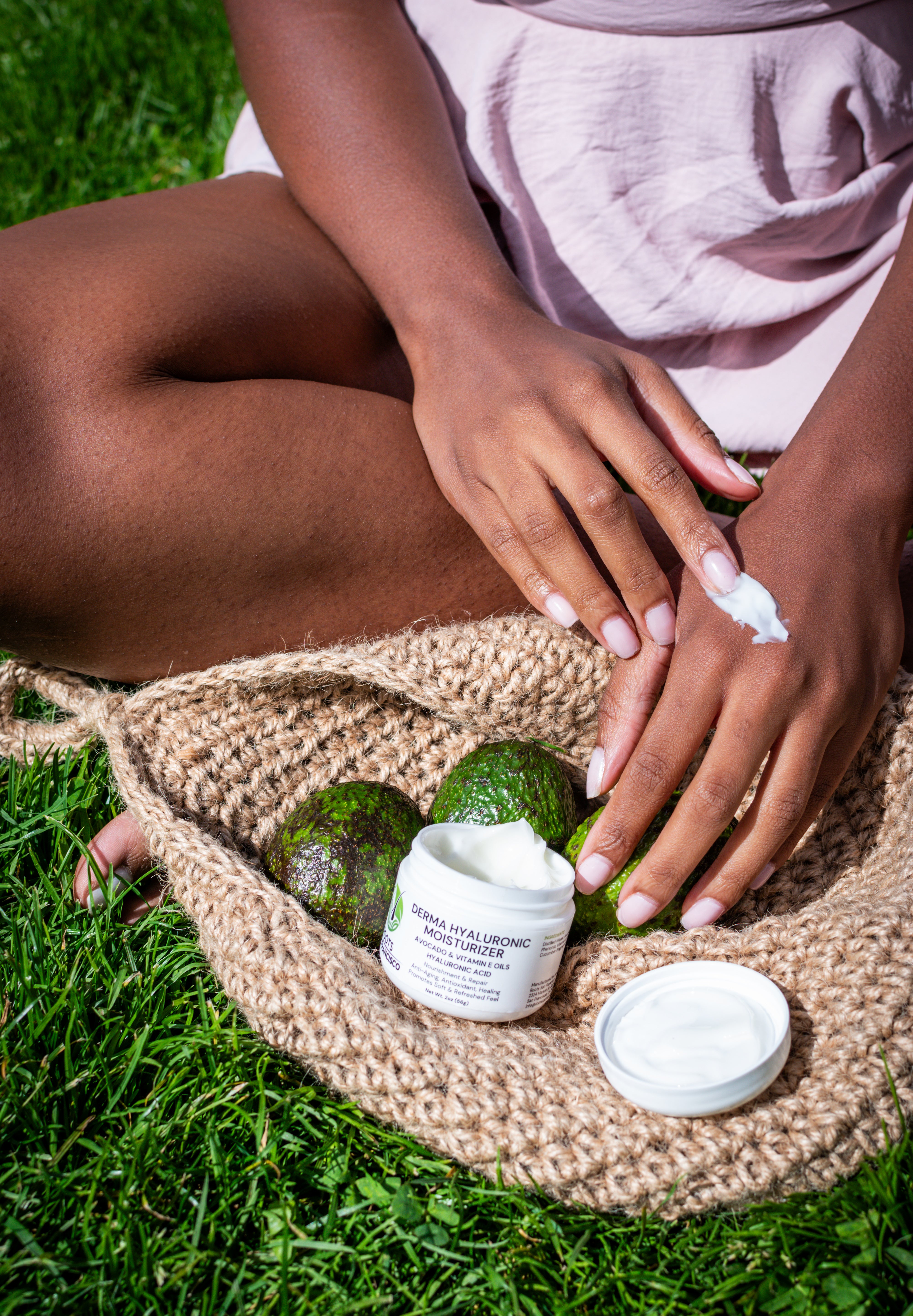 Person applying cream to hands with avocadoes and a jar of cream on a woven mat outdoors.
